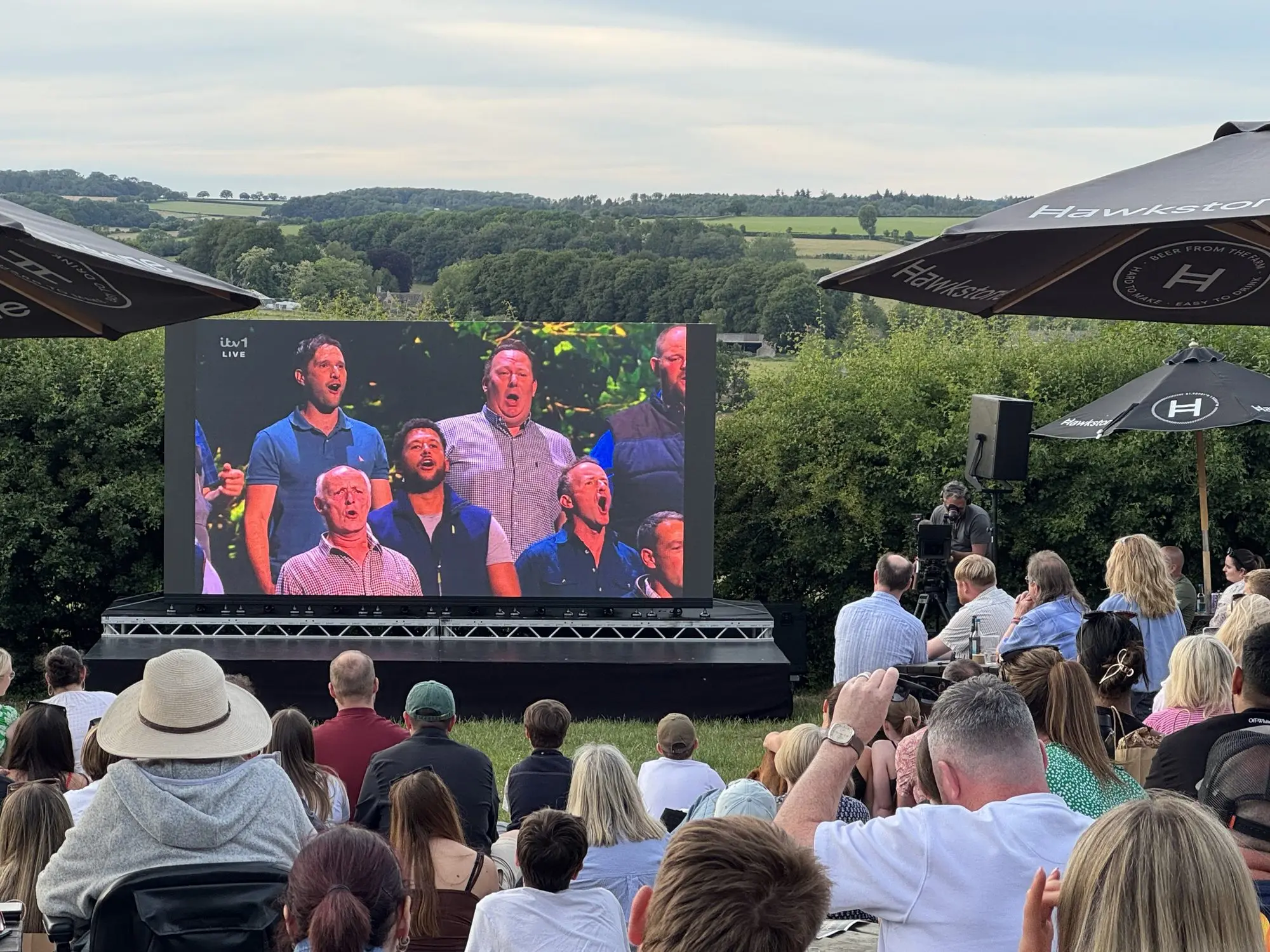 Crowd watching The Farmers Choir on a large outdoor LED screen with Cotswold hills behind