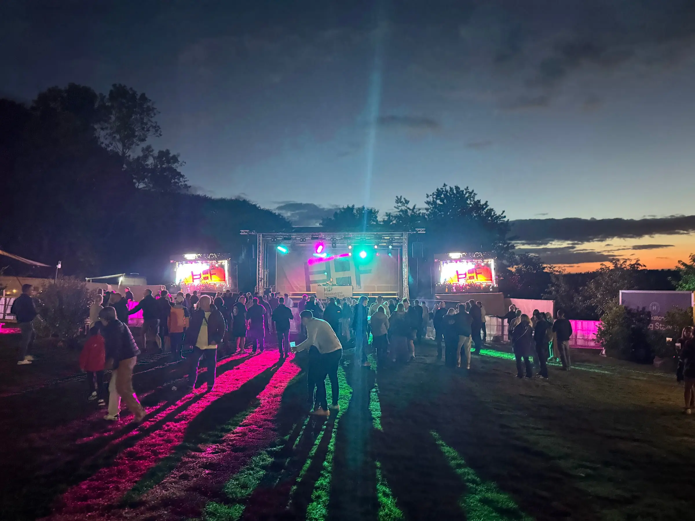 Colourful stage lighting and LED screens illuminating the crowd at dusk