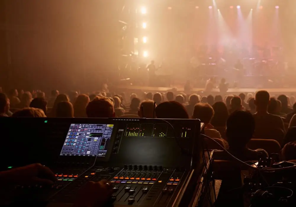 Sound engineer's hand on a mixing console with a lit stage and crowd in the background