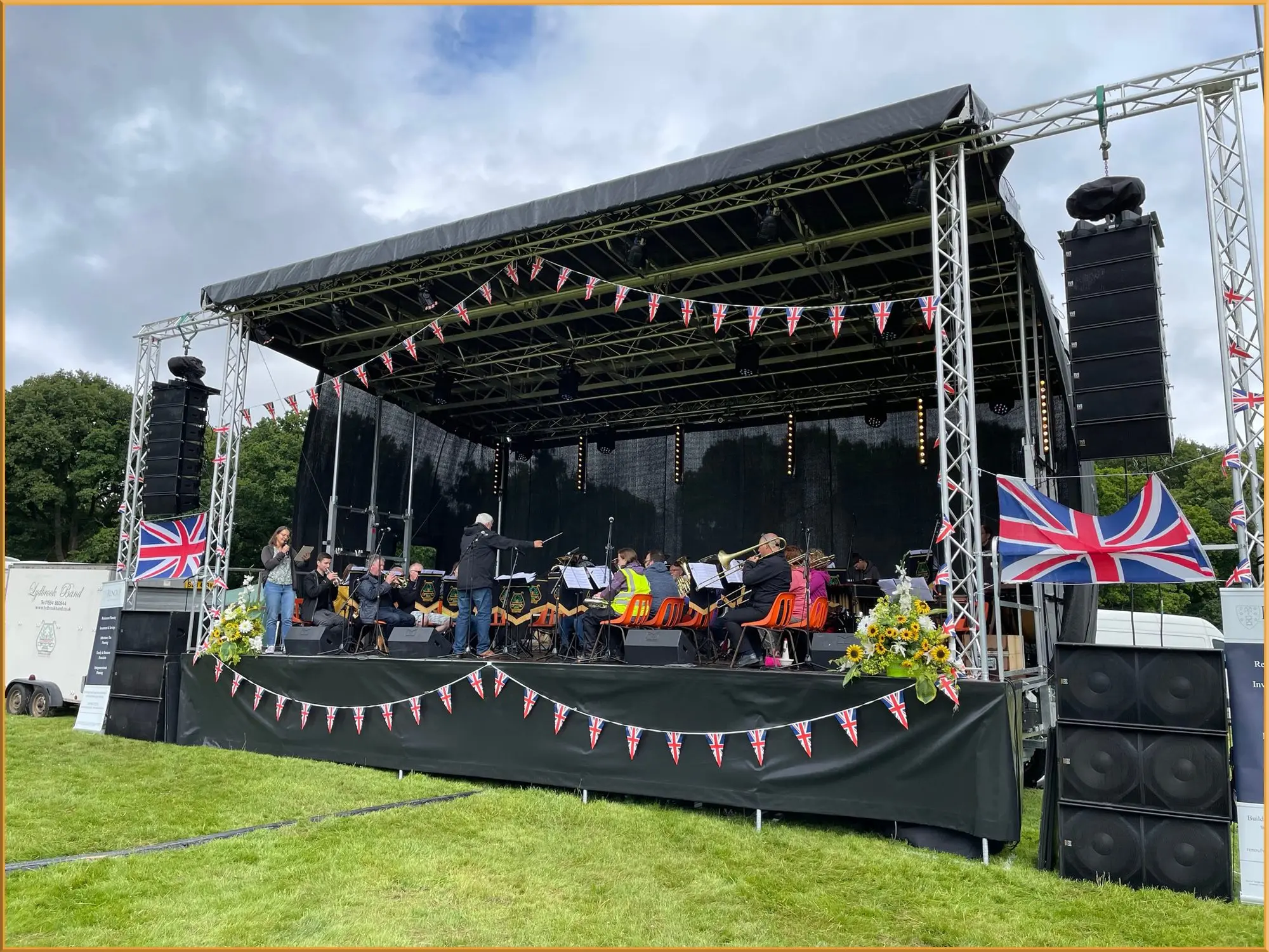 Outdoor stage decorated with bunting and Union Jack flags, with an orchestra performing
