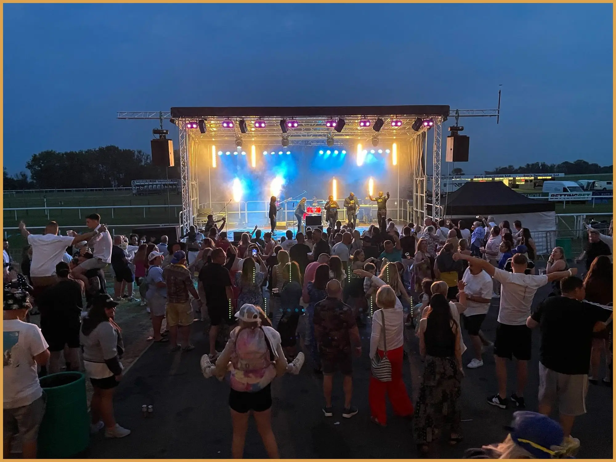 Crowd in front of a lit festival stage at Bath Racecourse at dusk