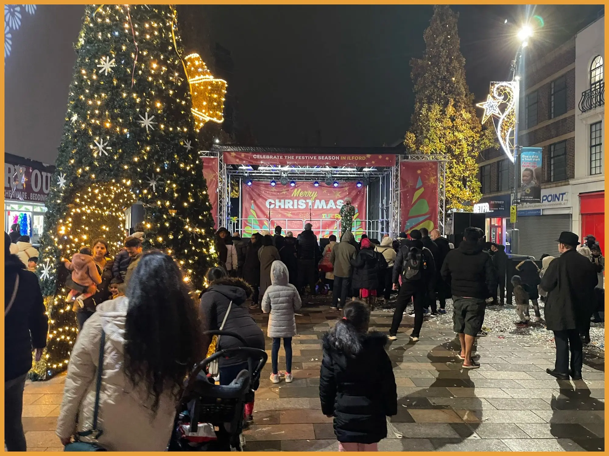 Crowd gathered around a lit Christmas tree in front of the stage on Ilford high street at night